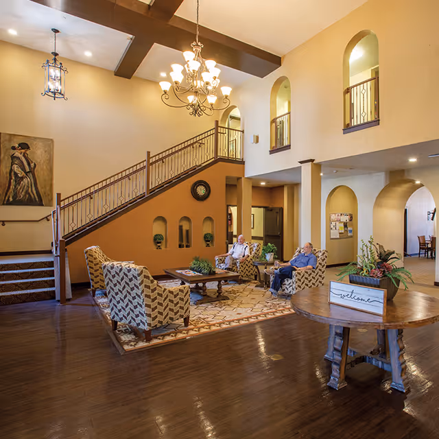 A spacious and warmly lit common area in an assisted living facility with high ceilings, a chandelier, and wooden beams. There are patterned armchairs arranged around a wooden coffee table on a decorative rug. Two elderly men are seated and conversing. A staircase with a decorative railing leads to an upper level with arched openings. A round wooden table with a welcome sign and a plant is in the foreground.