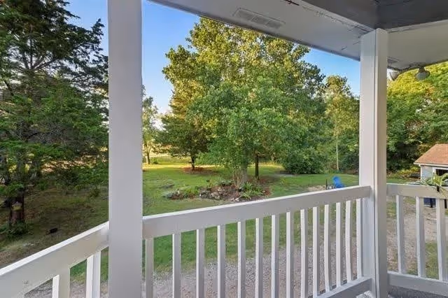 View from a covered porch with white railing overlooking a green yard with trees and a gravel path. There is a small structure with a brown roof visible to the right.