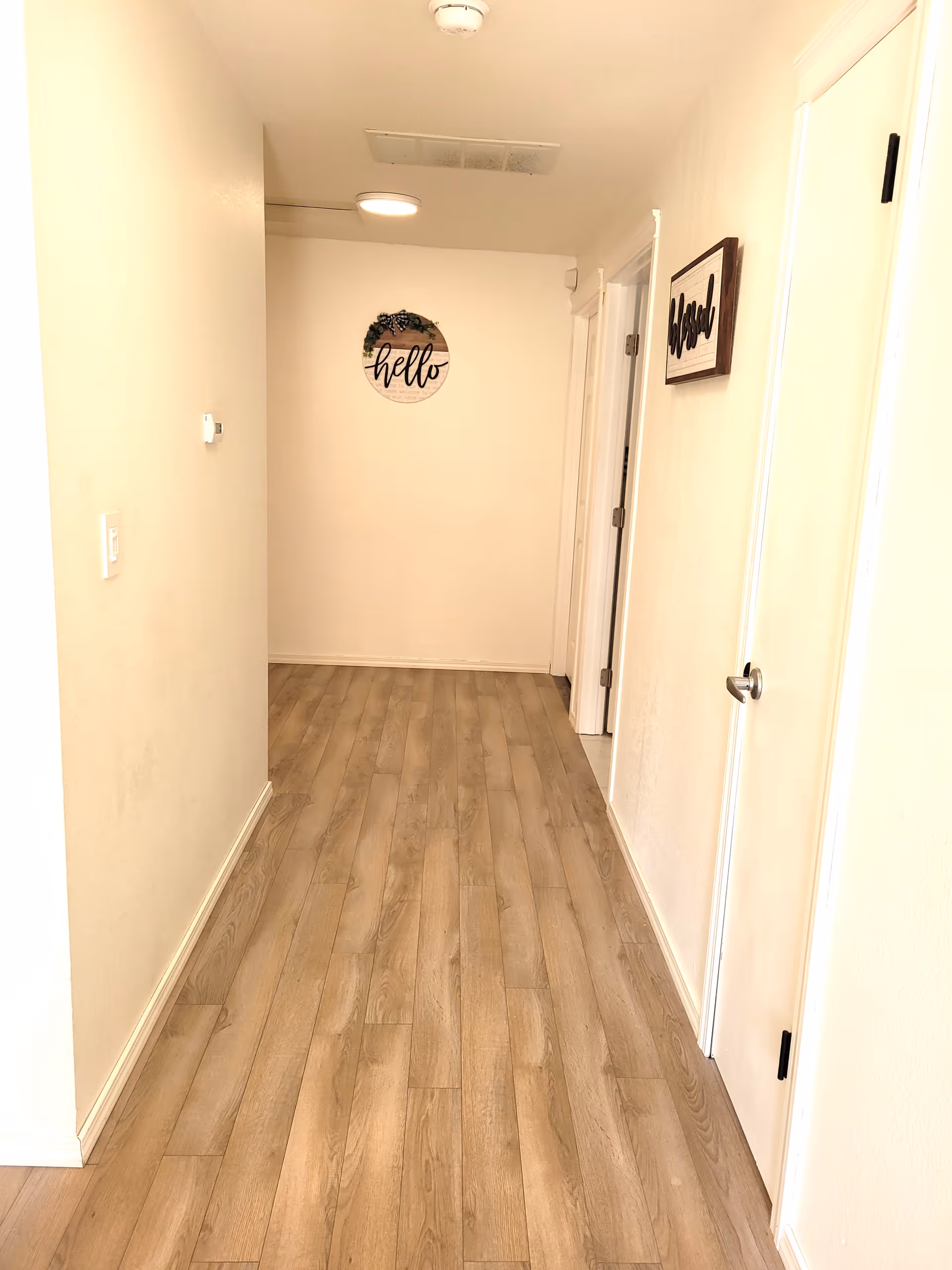 A hallway with light wood flooring and cream-colored walls. There are two white doors on the right side, one partially open. At the end of the hallway, a round wall decoration with the word 'hello' and some greenery is visible. Another rectangular wall decoration with the word 'bless' is hung on the right wall. The ceiling has a light fixture and an air vent.