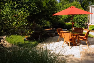 Sunny outdoor patio with a round wicker table, four chairs and a red umbrella surrounded by green landscaping.