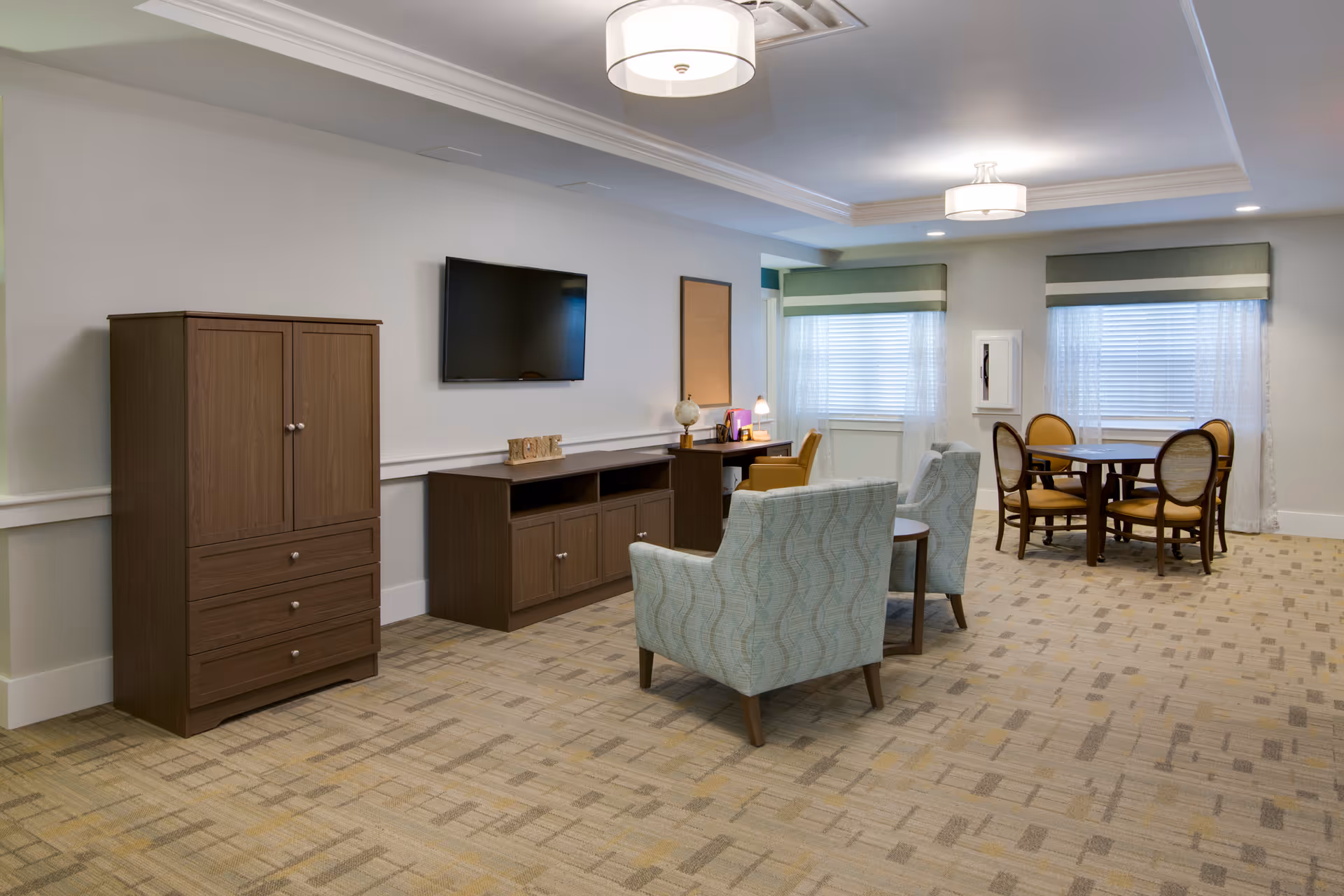 Bright communal lounge with upholstered armchairs, a wall-mounted TV and console, and a round dining table by windows.