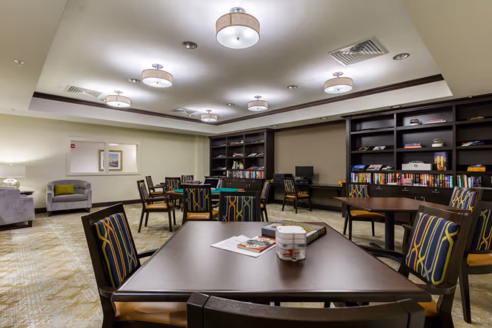 Well-lit communal room with multiple tables and patterned chairs, bookshelves, and lounge seating.