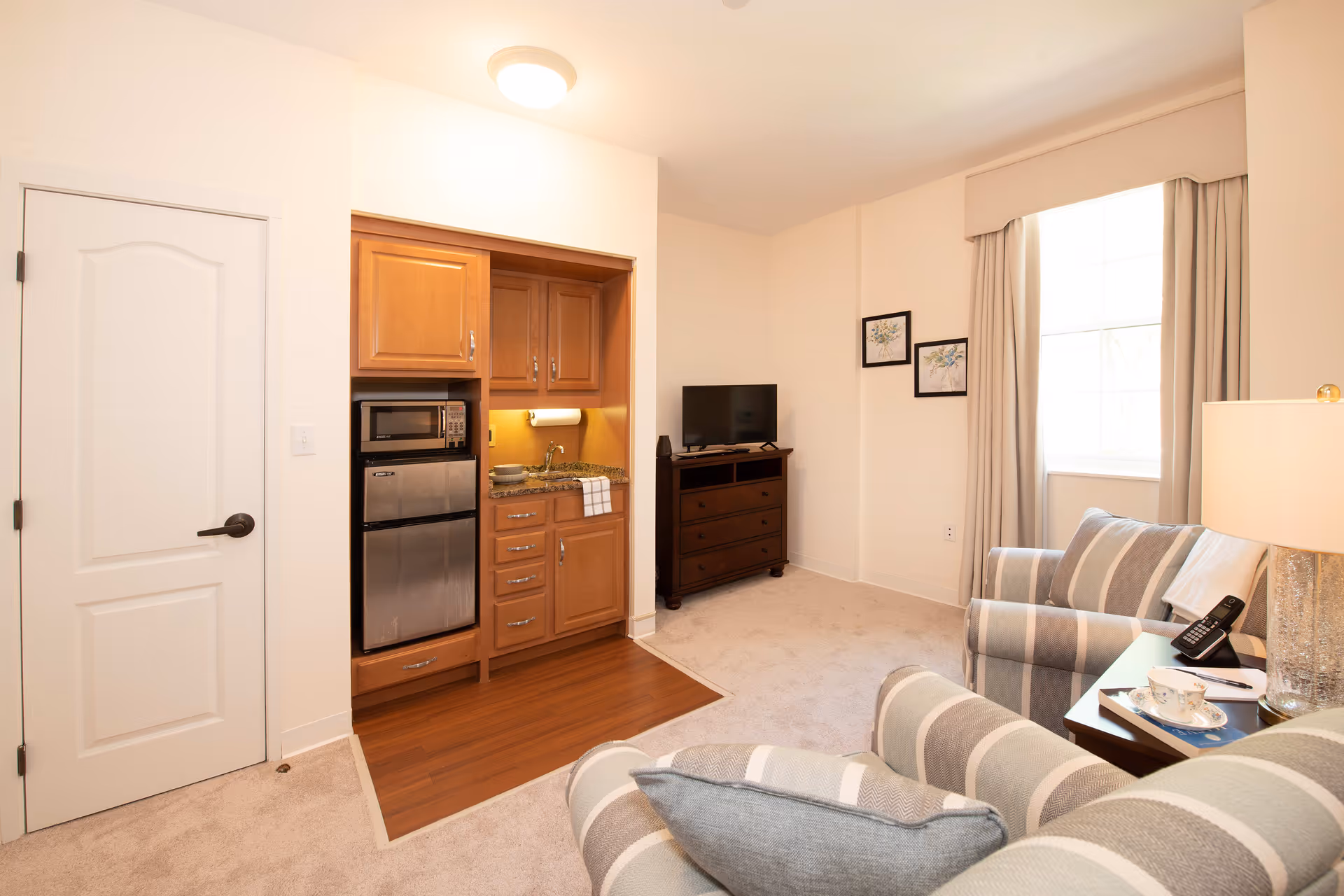 Bright living room with a small kitchenette, striped armchairs, a TV on a dresser, and a window with curtains.