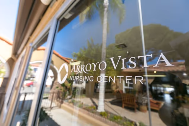 Glass door or window with the text 'Arroyo Vista Nursing Center' printed on it, reflecting an outdoor area with palm trees, a bench, and a building with a tiled roof.