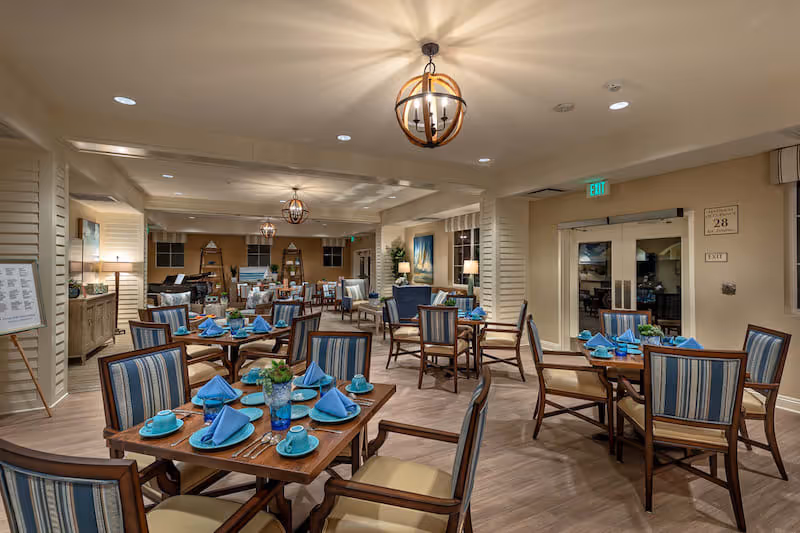 Bright communal dining room with multiple wooden tables set with blue napkins and dishware, surrounded by upholstered chairs and decorative pendant lighting.