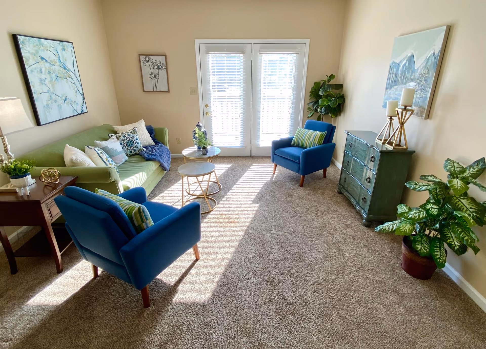A bright living room with beige walls and carpeted floor. The room features a green sofa with multiple decorative pillows, two blue armchairs with striped cushions, two round nesting coffee tables, a wooden side table with a lamp and plant, a green chest of drawers with three candles on top, and two framed paintings on the walls. Sunlight streams through white blinds covering French doors at the back, and there is a large potted plant in the corner.
