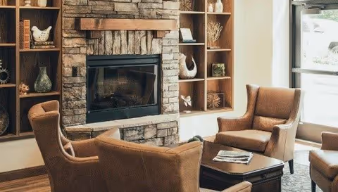 A cozy living room area with two brown upholstered armchairs and a matching sofa arranged around a dark wooden coffee table. Behind the seating is a stone fireplace with built-in wooden shelves on either side, decorated with vases and small sculptures. Natural light comes in from a glass door on the right side.