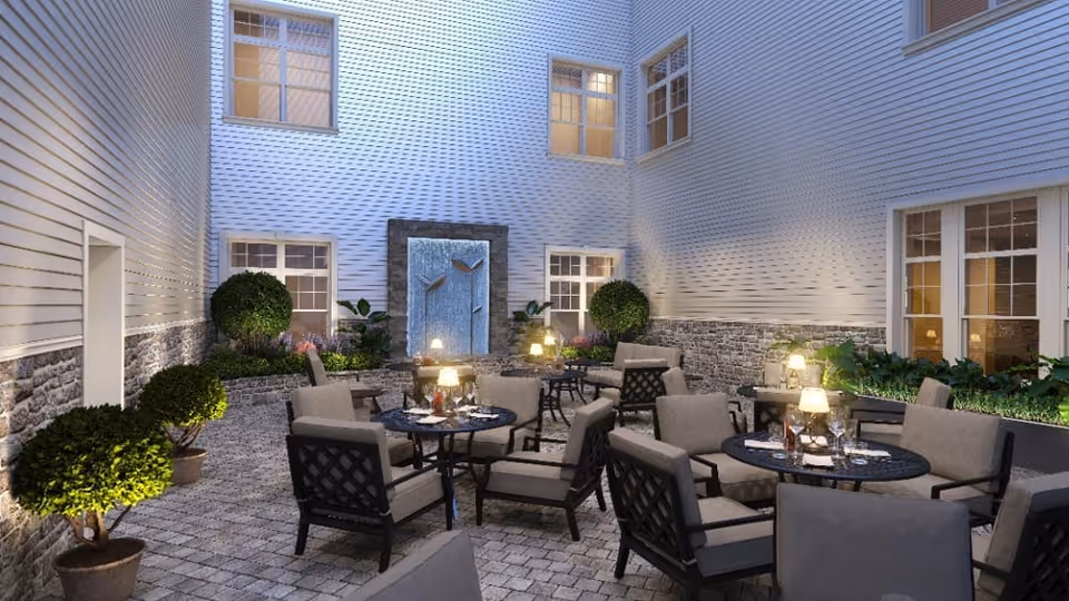 Outdoor courtyard area with multiple round tables and cushioned chairs arranged for seating. The space is surrounded by building walls with windows and decorated with potted plants and shrubs. A water feature with a stone frame and metal leaf decorations is visible on one wall, illuminated by soft lighting.