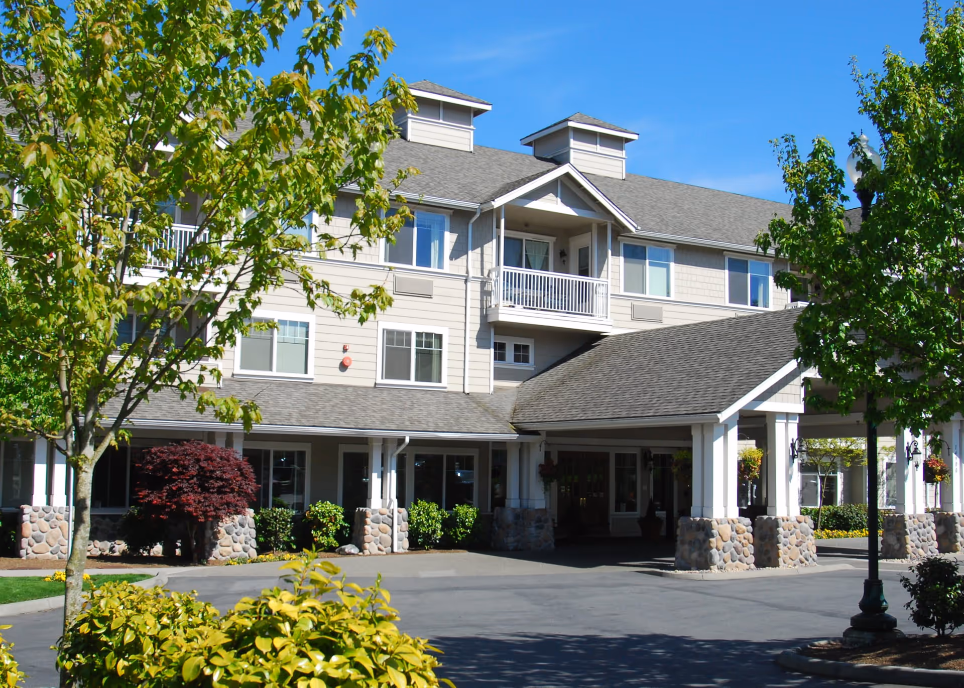 Exterior view of a multi-story senior living facility building with a covered entrance supported by stone pillars, surrounded by green trees and shrubs under a clear blue sky.