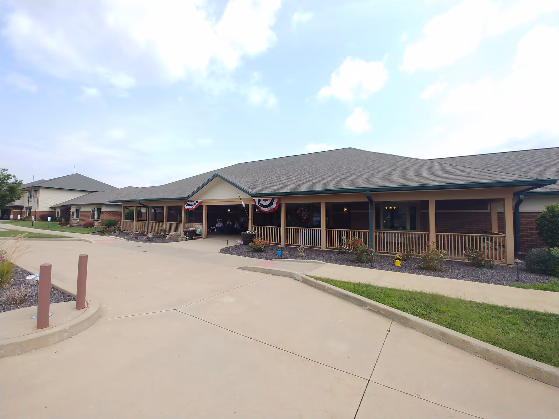 Exterior view of a single-story building with a covered porch decorated with red, white, and blue bunting. The building has a gray shingled roof, beige siding with brick accents, and a paved driveway leading up to the entrance. There are landscaped areas with bushes and flowers along the front.