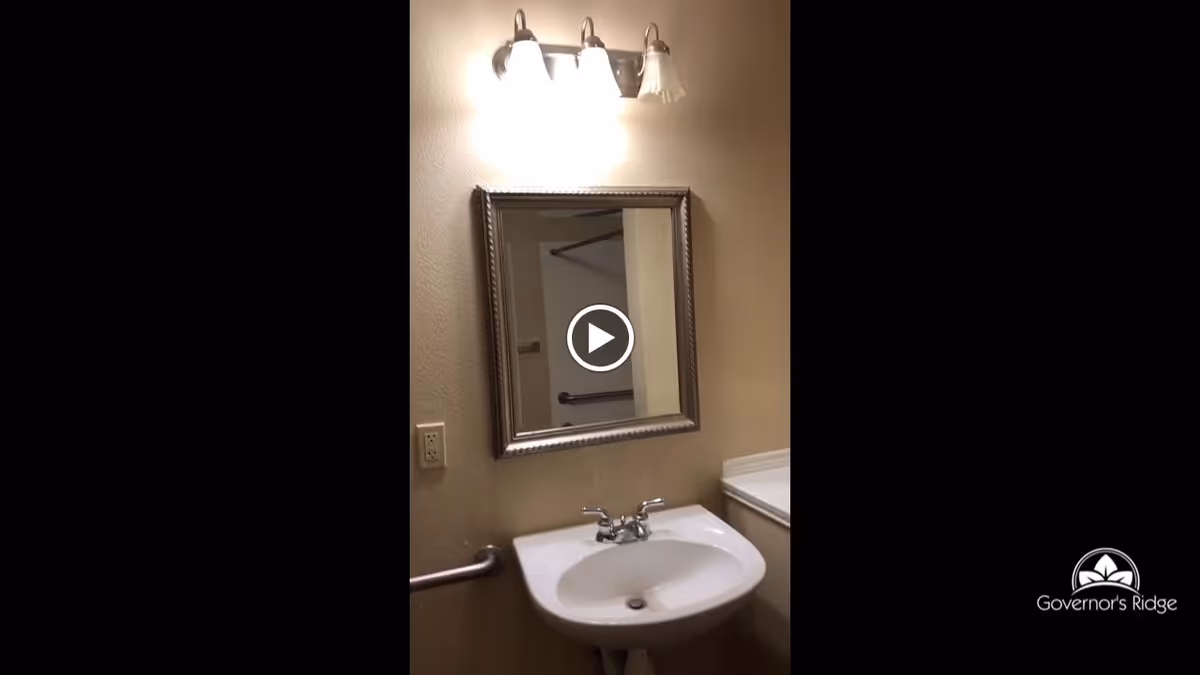 A bathroom sink with a silver faucet beneath a rectangular mirror with a decorative frame. Above the mirror is a three-light fixture with frosted glass shades. A grab bar is mounted on the wall to the left of the sink, and a countertop is visible to the right. The wall is painted beige.