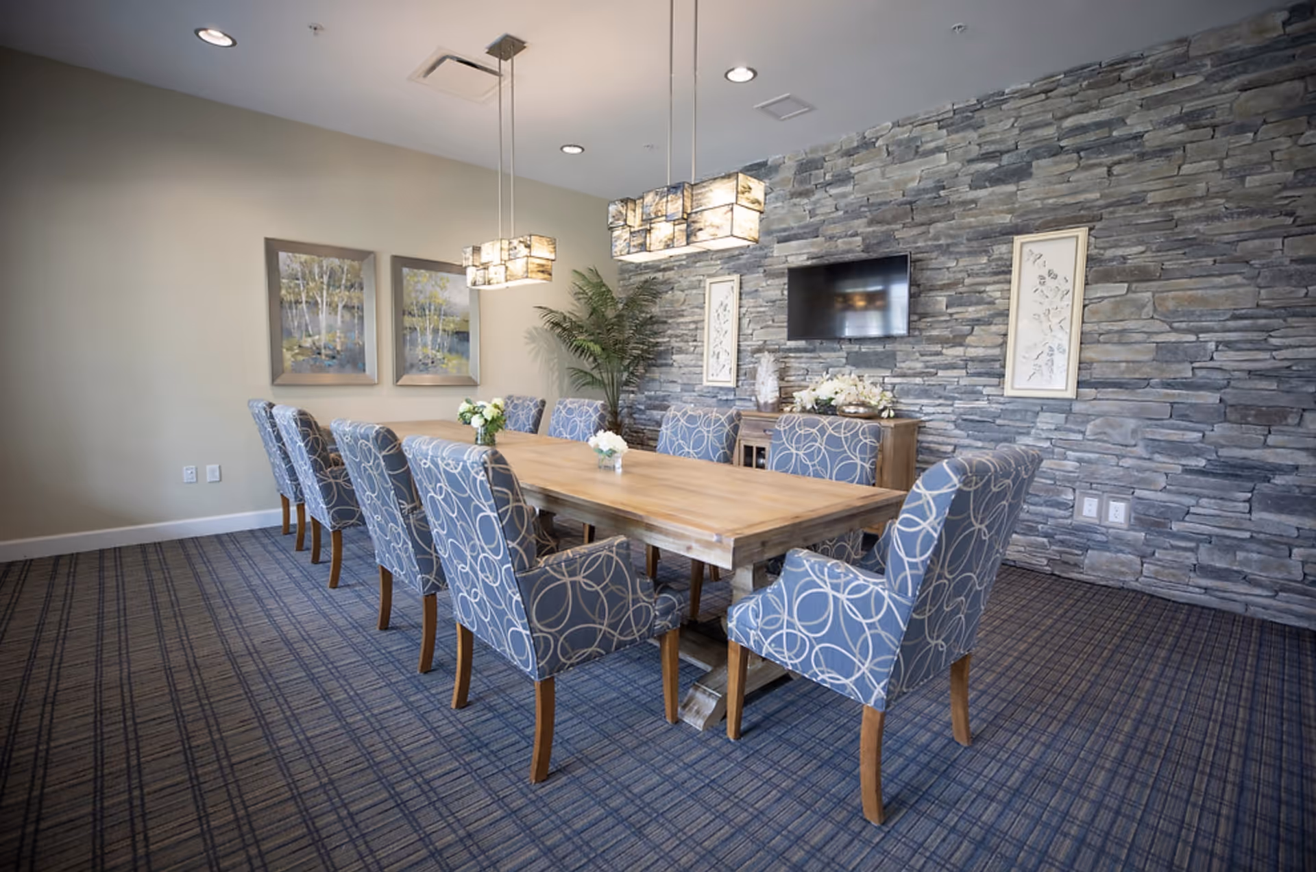 A dining room with a long wooden table surrounded by ten blue upholstered chairs with circular patterns. The room features a stone accent wall with a mounted flat-screen TV, two framed artworks, and decorative plants. Two modern rectangular pendant lights hang above the table, and the floor is covered with a blue patterned carpet.