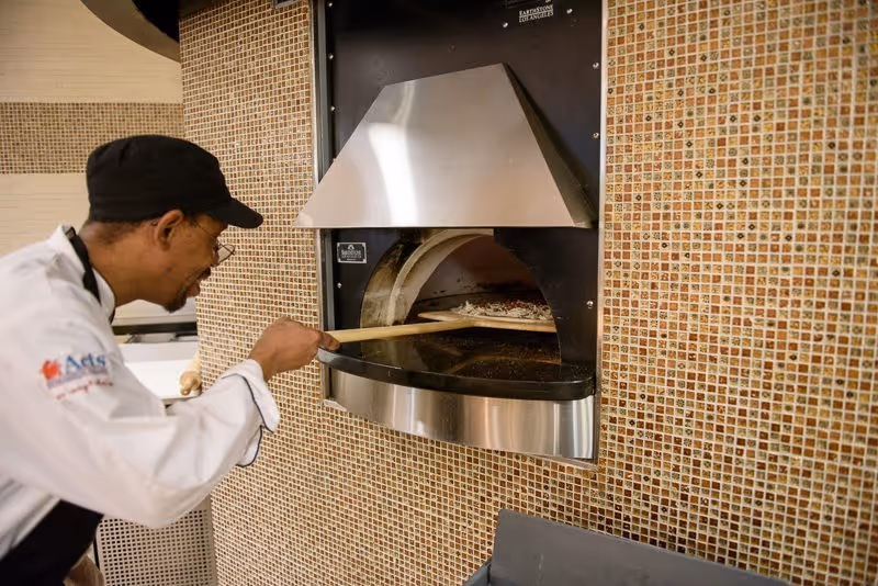 A chef wearing a black cap and white uniform is placing a pizza into a built-in stone oven with a wooden pizza peel. The oven is set into a wall covered with small square mosaic tiles in shades of brown and beige.