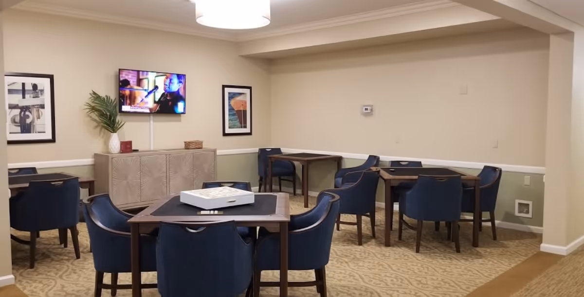 A common area with several square tables surrounded by blue upholstered chairs. A TV is mounted on the wall above a decorative cabinet, and two framed pictures hang on the wall. The room has beige walls with a green lower half and patterned carpet flooring.