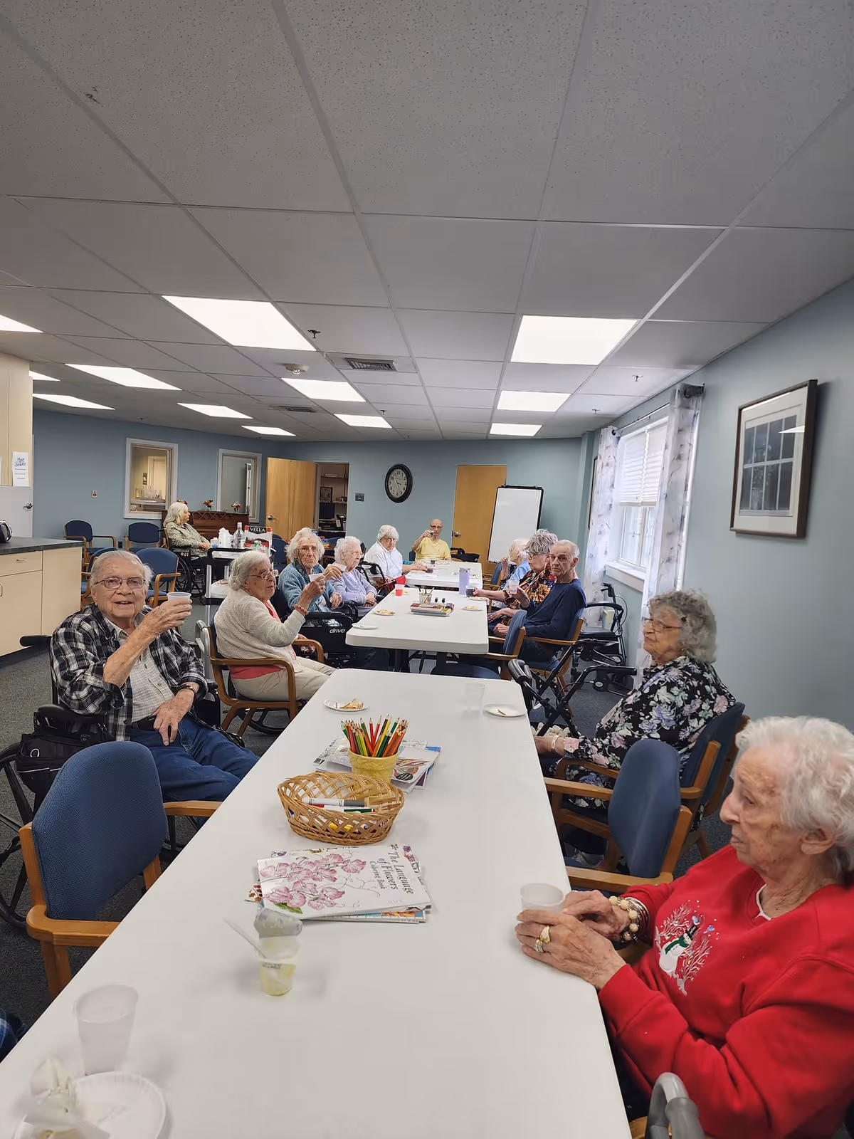 A group of elderly people sitting around two long tables in a well-lit room with light blue walls and a white ceiling. Some are holding cups and appear to be socializing. There are colored pencils in a basket on the table along with some papers and plates. The room has windows with curtains, a clock on the wall, and a whiteboard in the background.