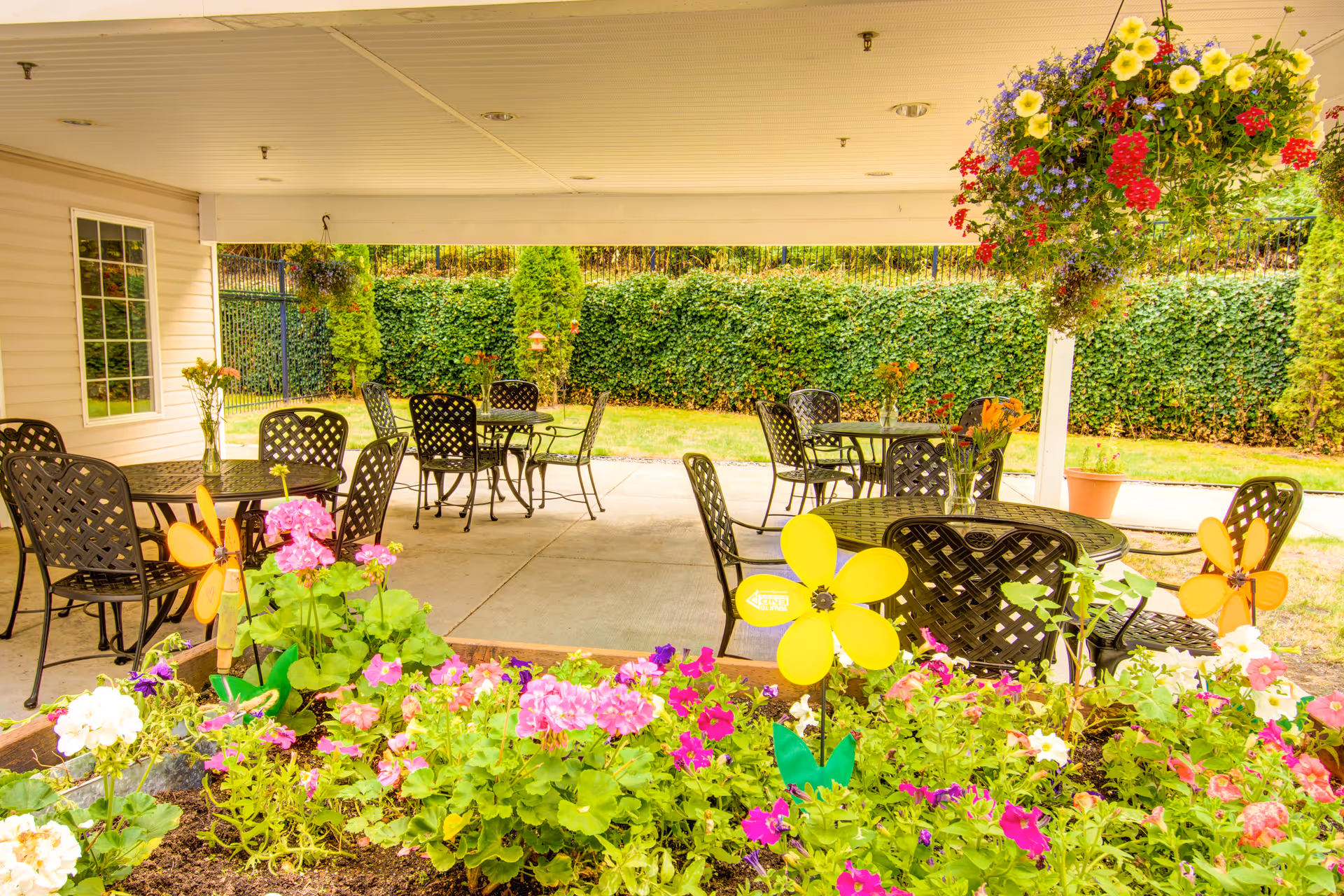 Covered outdoor patio area with several black metal tables and chairs, surrounded by colorful flowers and greenery, including hanging flower baskets and garden decorations shaped like yellow and orange flowers.
