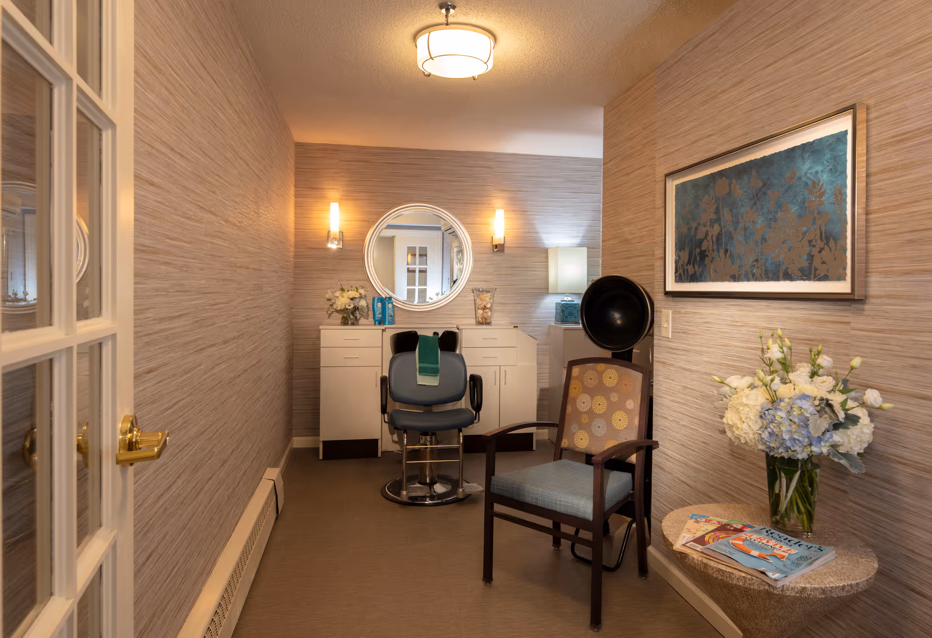 A small salon room with a styling chair in front of a white cabinet and a round mirror on the wall. The room has beige textured walls, a ceiling light, and two wall sconces. There is a second chair with a patterned cushion next to a small round table holding a vase of white and blue flowers and some magazines. A framed artwork hangs on the wall above the table.