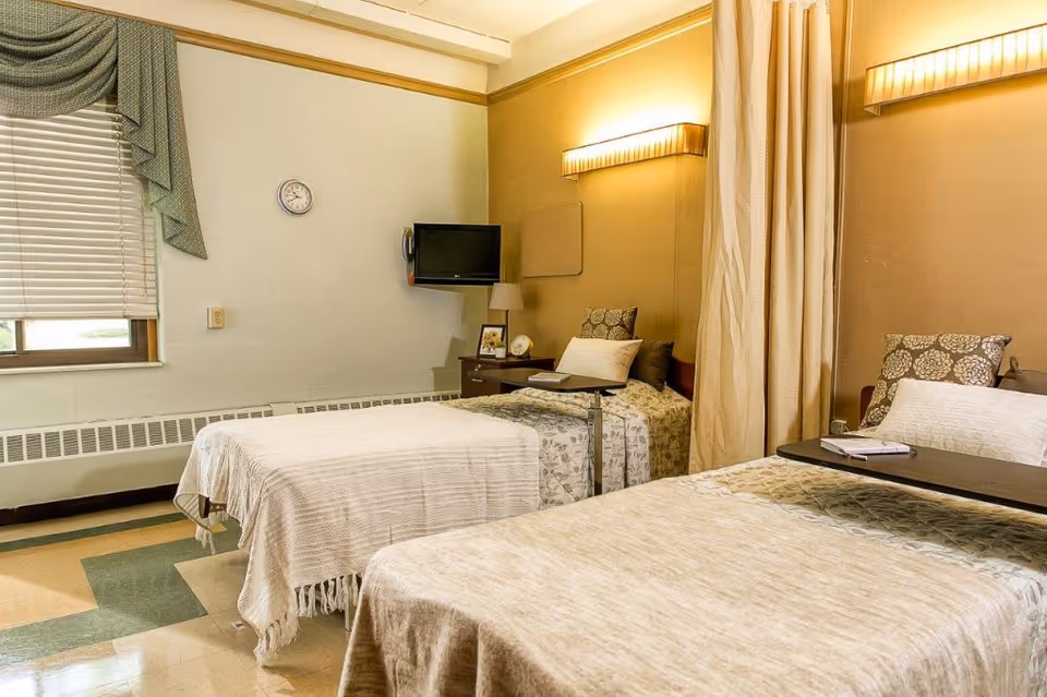Two twin beds in a warmly lit senior living bedroom with bedside tables, a wall-mounted TV, and a curtained divider.