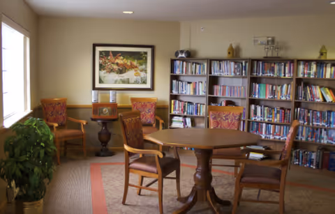 A cozy room with a wooden octagonal table surrounded by four wooden chairs with red patterned cushions. Behind the table, there are three bookshelves filled with books. On the wall above the bookshelves, there is a framed picture of a floral arrangement. To the left, there are two additional chairs with similar red patterned cushions and a small round table with a vintage radio on it. A large window on the left side lets in natural light, and a green potted plant is placed near the window.