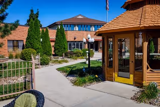 Outdoor view of a senior living facility named St Francis Terrace featuring a paved walkway, green shrubs, tall trees, a wooden gazebo, and a brick building with multiple windows under a clear blue sky.