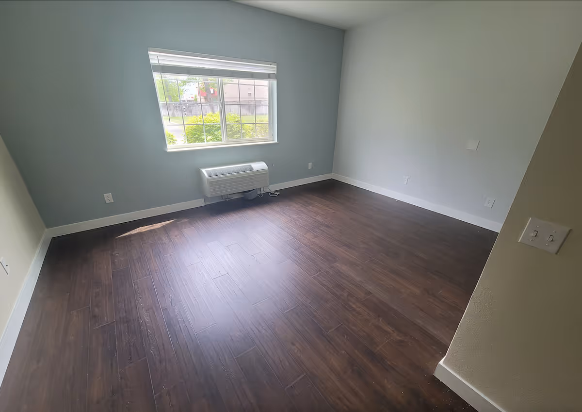 Empty room with dark wood flooring, light gray walls, a large window with white blinds, and a wall-mounted air conditioning unit below the window.