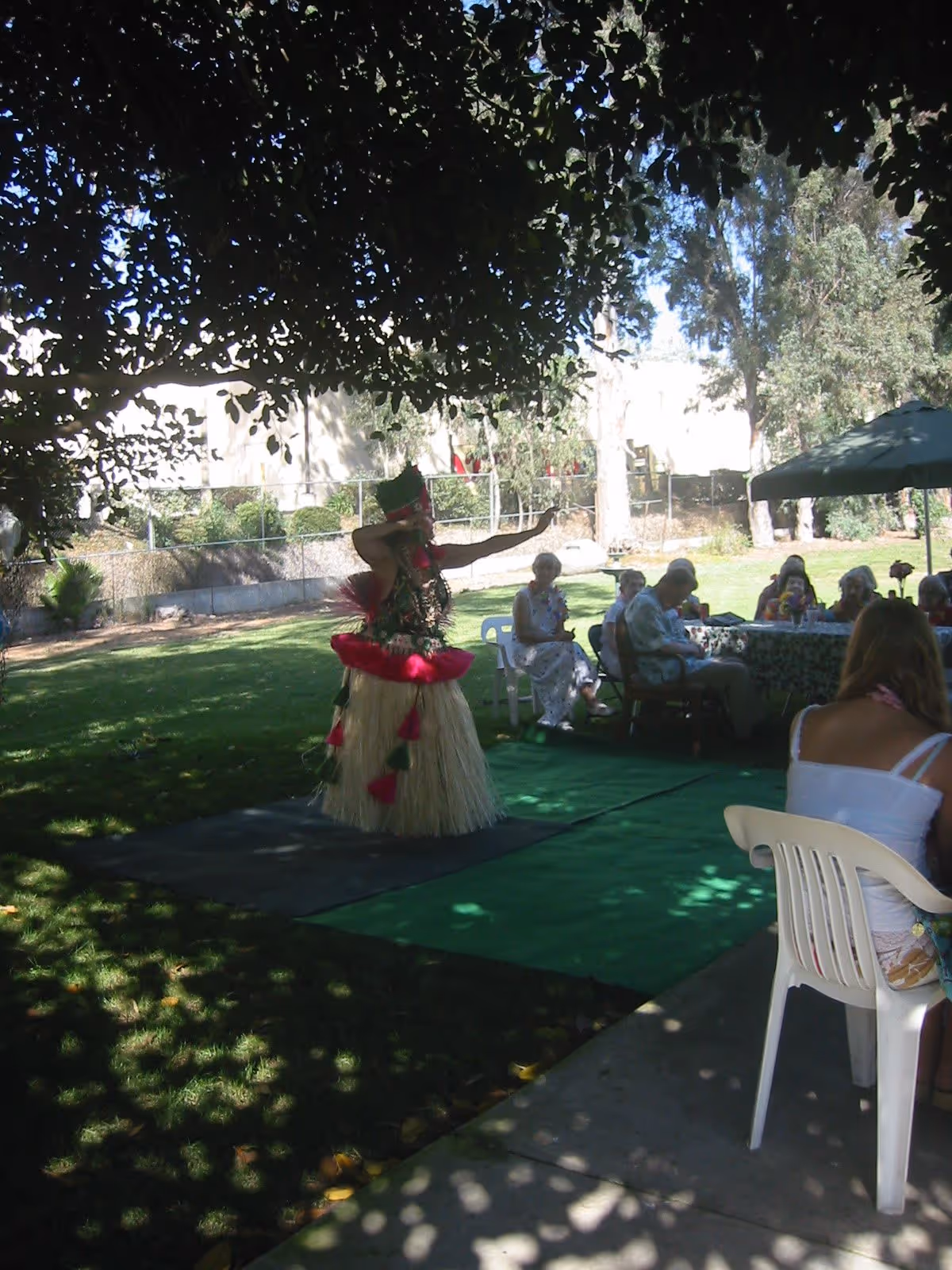 An outdoor scene at Shadowridge Senior Living showing a person dressed in traditional Hawaiian attire performing a hula dance on a green mat. Several elderly people are seated around a table under an umbrella, watching the performance. The area is shaded by trees with sunlight filtering through the leaves.