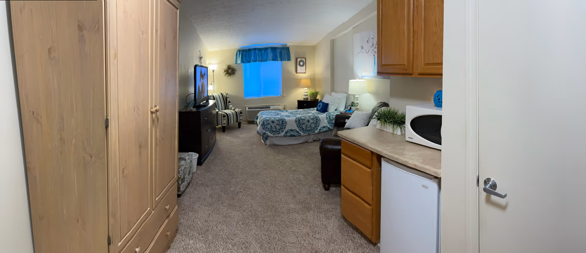Interior view of a senior living facility room at The Woodlands of Columbus featuring a bed with blue and white bedding, a TV on a dresser, a striped armchair, a small kitchenette area with a microwave and mini fridge, wooden cabinets, a window with blue curtains, and carpeted floor.