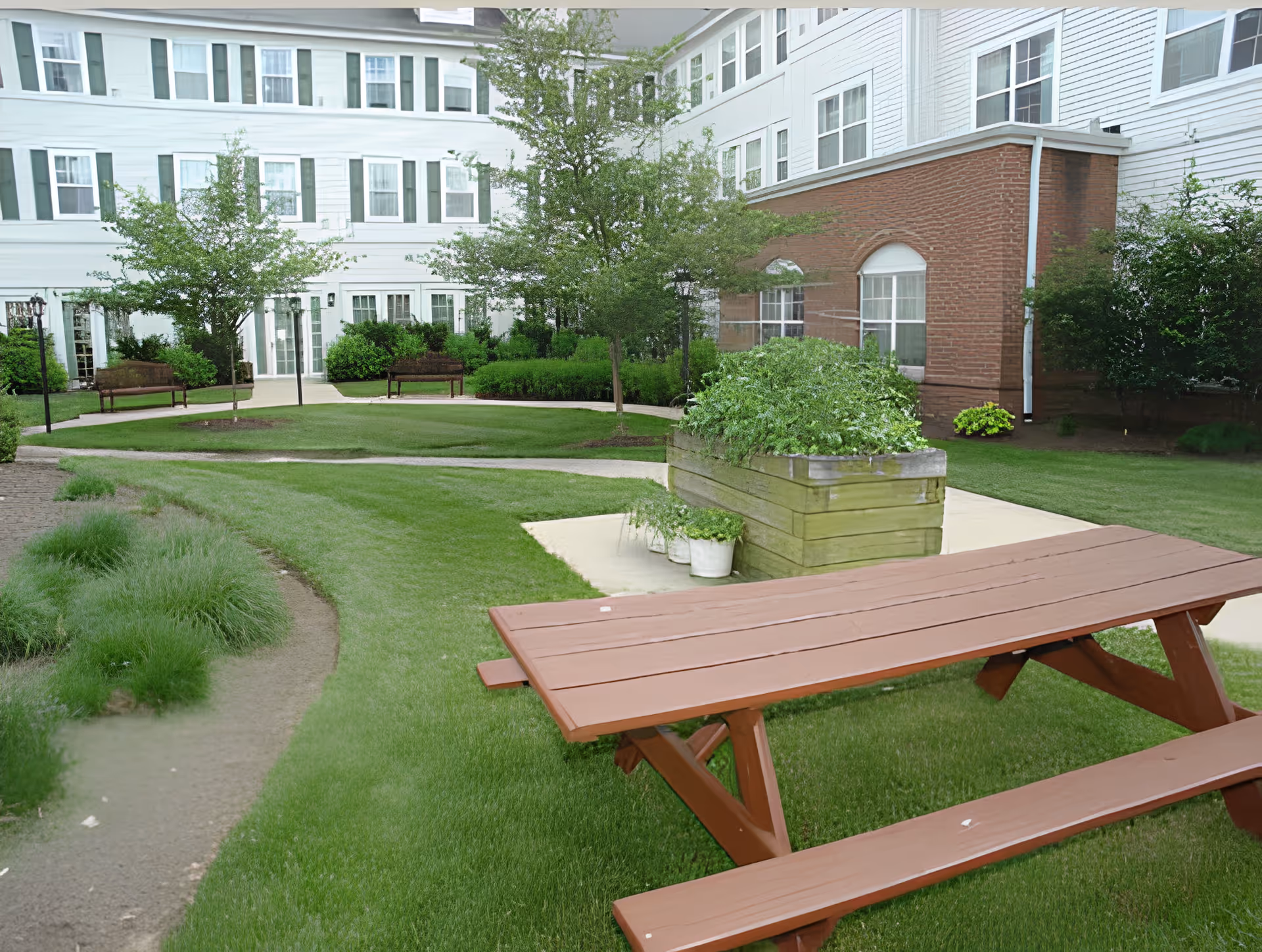 A landscaped courtyard with a wooden picnic table, raised planter, benches and the facade of a multi-story senior living building.
