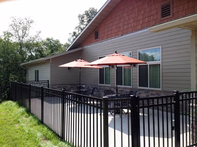 Outdoor patio area with black metal tables and chairs under two red umbrellas, surrounded by a black metal fence. The patio is adjacent to a beige building with stone accents and several windows. Trees and greenery are visible in the background.