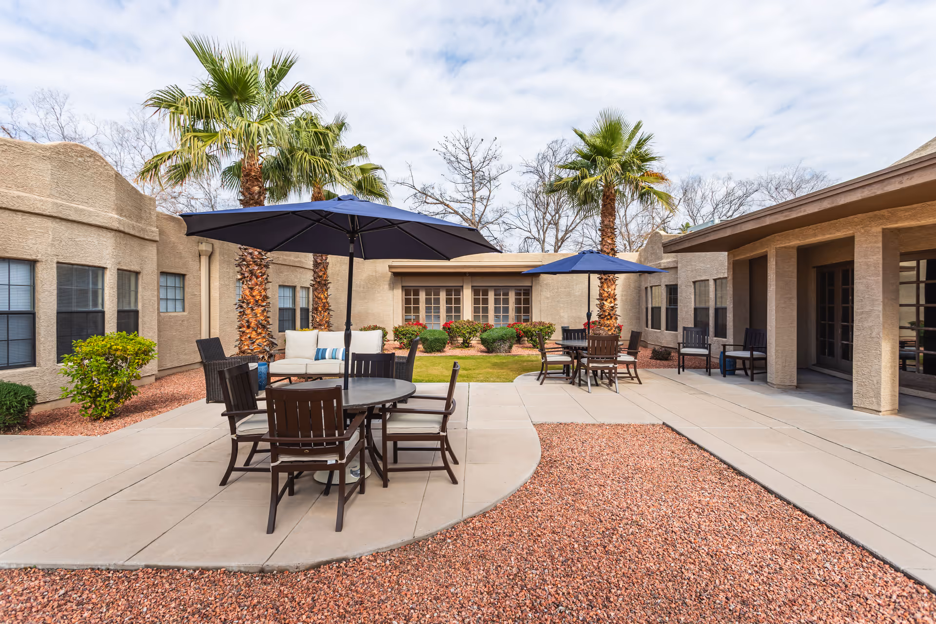 Outdoor patio area at a senior living facility with round tables, chairs, and large blue umbrellas. The patio is surrounded by beige stucco buildings, palm trees, and landscaped bushes under a partly cloudy sky.