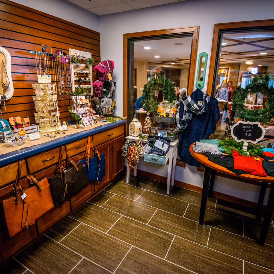 Interior view of a small retail shop area within a senior living facility, displaying various items for sale including handbags hanging on wooden cabinets, jewelry on a countertop, scarves, wreaths, and specialty tunics on a round table. Two large windows show people and more decorations in an adjacent room.