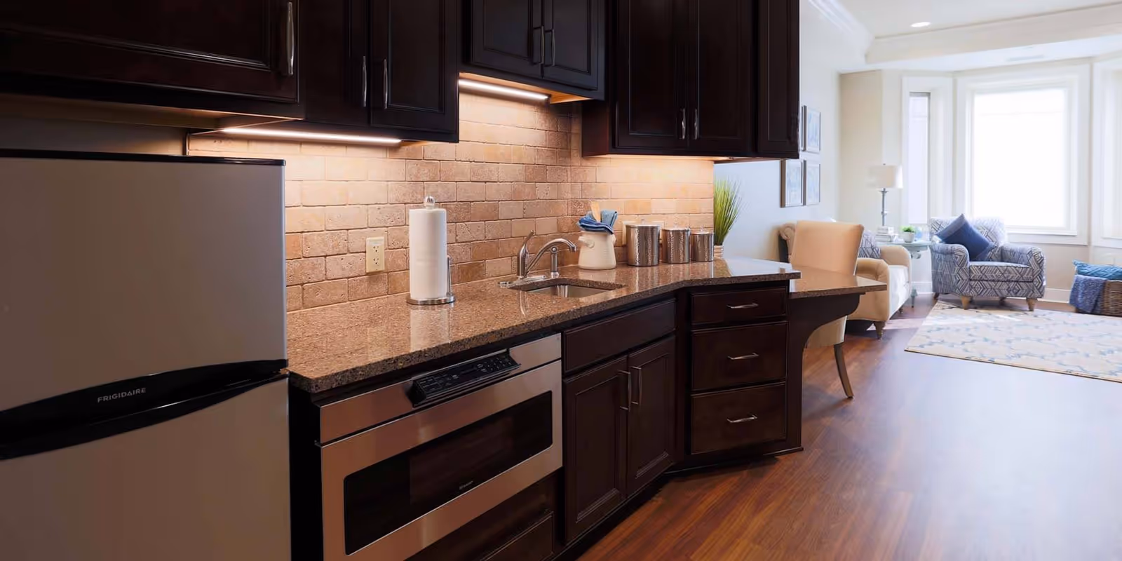 Open kitchen with dark wood cabinets, granite countertop, stainless steel refrigerator and microwave, and a sink that opens into a bright living room area.