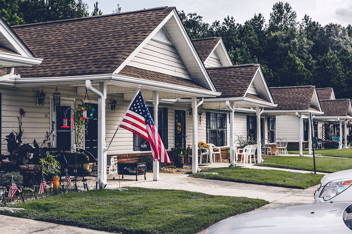 Row of single-story cottages with porches, American flags, and manicured lawns in front.