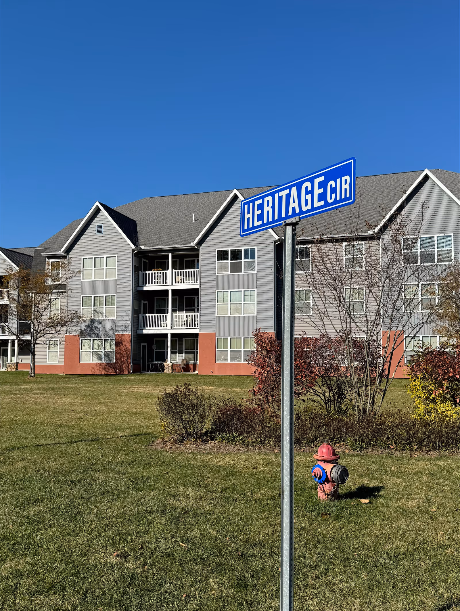 Exterior view of a multi-story residential building with balconies, a 'HERITAGE CIR' street sign in the foreground and a fire hydrant on a grassy lawn.