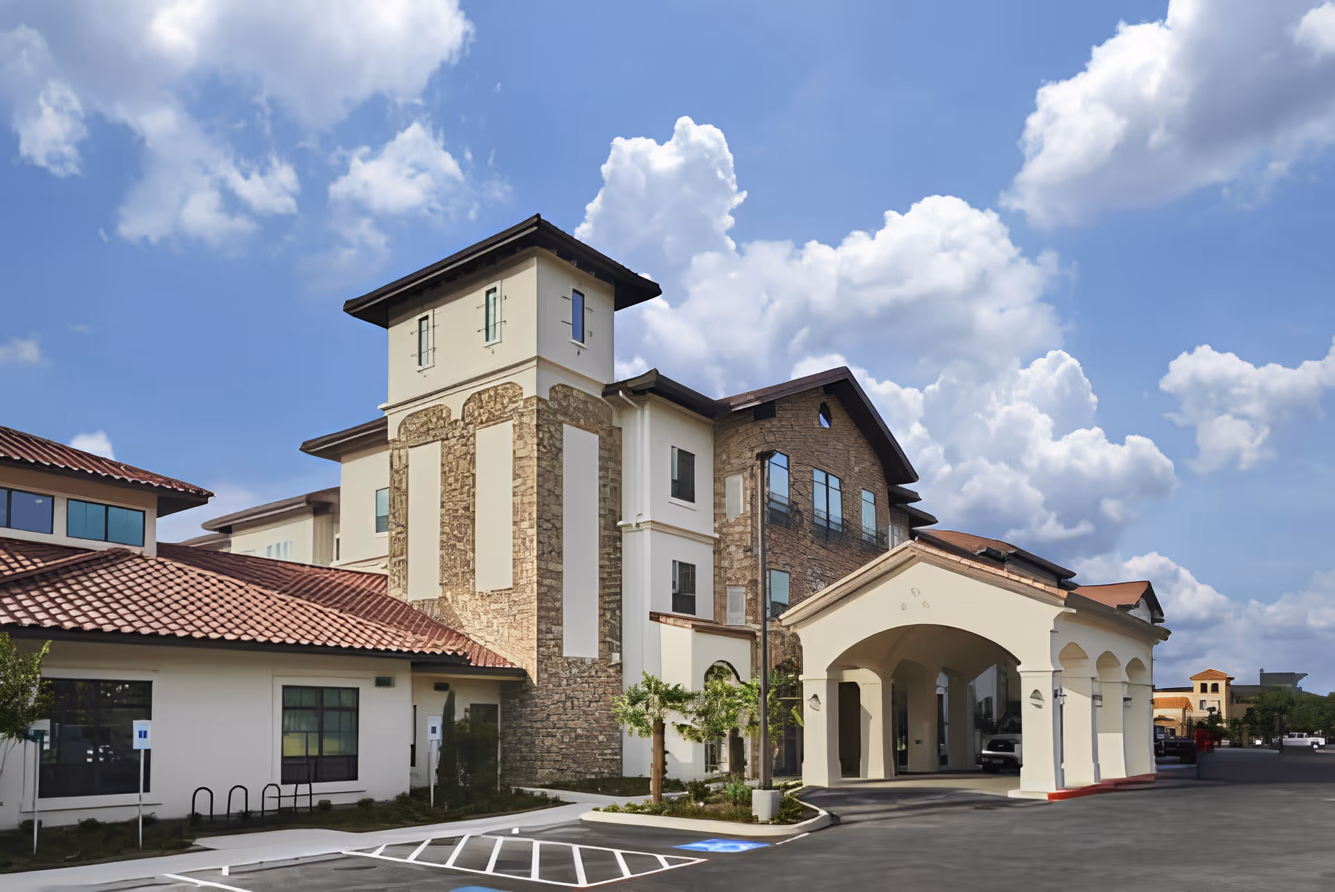 Exterior view of a multi-story assisted living and memory care facility with a stone and stucco facade, red tile roof, and covered entrance driveway under a partly cloudy blue sky.