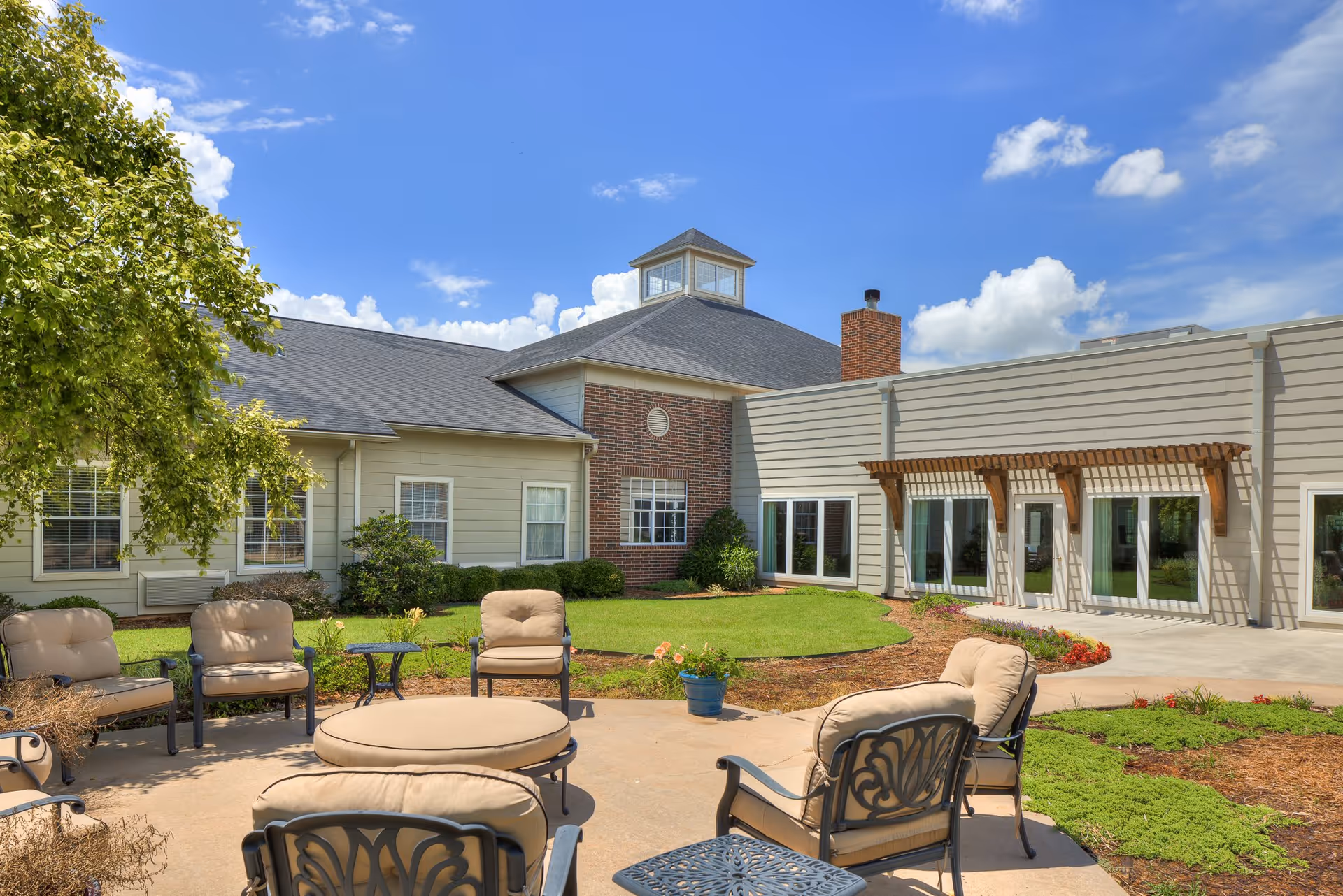 Outdoor patio area with cushioned chairs and a round ottoman arranged around a small table, surrounded by a well-maintained garden and lawn. The building in the background has beige siding, brick accents, multiple windows, and a pergola above a set of glass doors under a bright blue sky with scattered clouds.