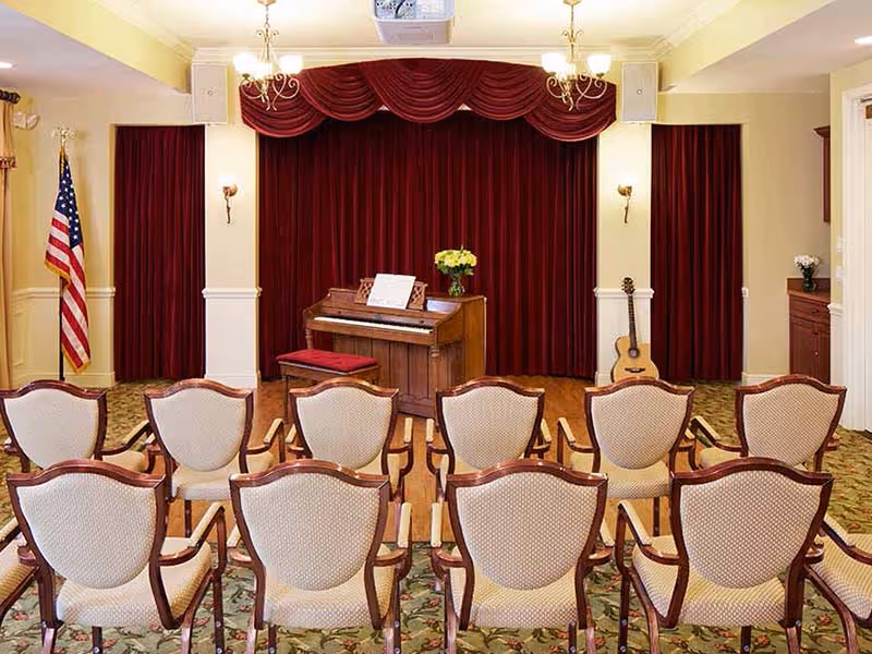 A small auditorium or performance room with rows of upholstered chairs facing a wooden piano on a stage with red velvet curtains. An American flag stands to the left, and a guitar rests against the wall on the right. The room has chandeliers and wall sconces providing warm lighting.