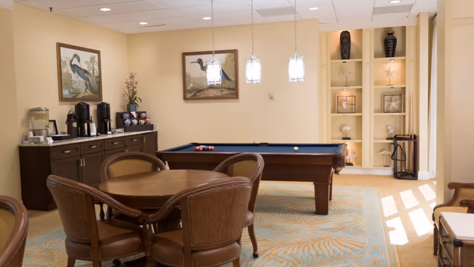 A recreational room with a pool table, a round wooden table with four chairs, and a sideboard with coffee and water dispensers. The room has beige walls, two framed bird paintings, built-in shelves with decorative items, and three hanging pendant lights above the pool table.