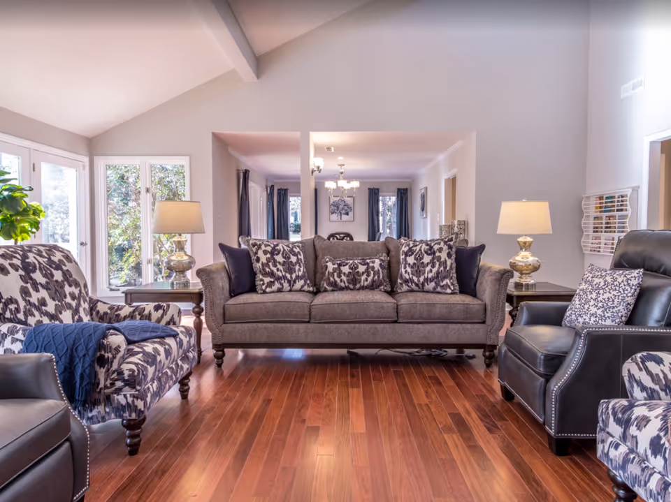 Bright living room with a gray upholstered sofa flanked by patterned armchairs, side tables with lamps, and hardwood floors.
