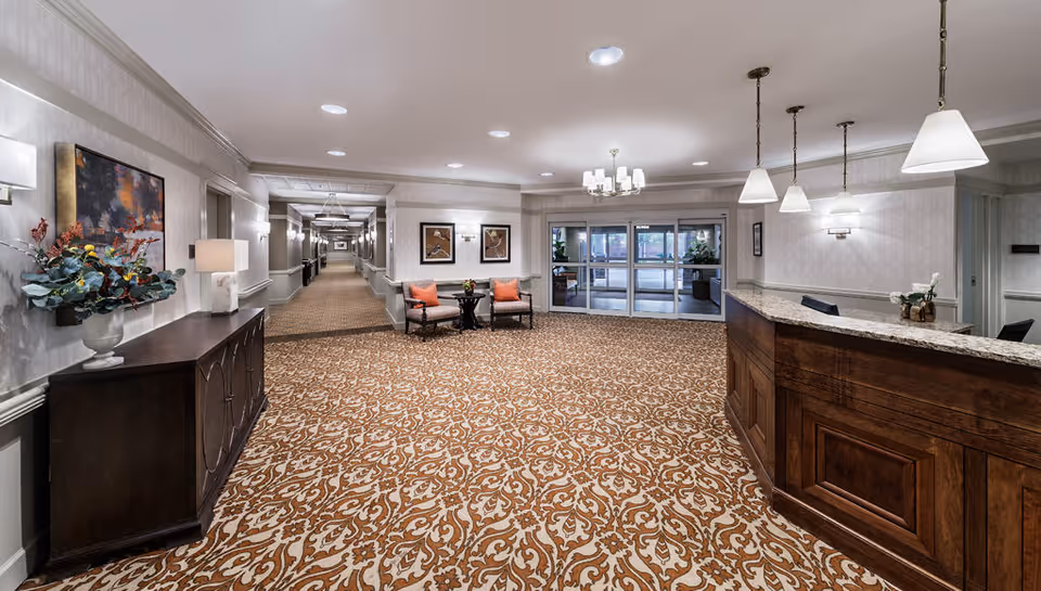 Spacious carpeted lobby with a wooden reception desk, seating area, decorative console table, and double glass doors at the far end.