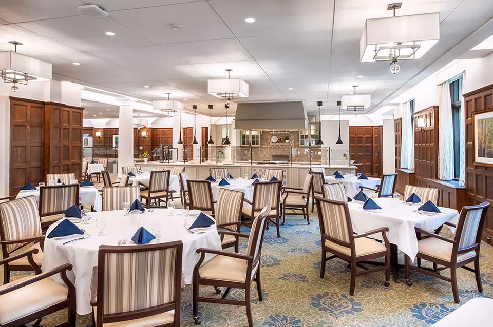 A spacious dining room with multiple round and square tables covered with white tablecloths and set with blue folded napkins, glasses, and silverware. The room features wooden paneled walls, large windows with curtains, patterned carpet, and modern ceiling lights. In the background, there is a kitchen or serving area with a large hood and hanging pendant lights.