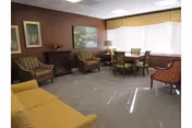 A cozy living room area with a yellow sofa, two striped armchairs, a wooden table with four chairs near a large window with blinds, and framed artwork on the walls. The room has a carpeted floor and soft lighting.