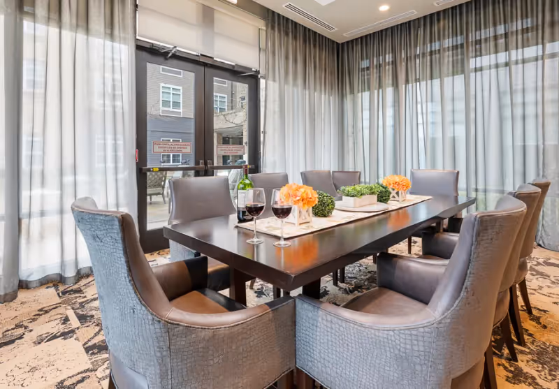 Bright dining/meeting room with a long dark wooden table surrounded by upholstered chairs, wine glasses and floral centerpieces, and sheer curtains covering glass doors in the background.
