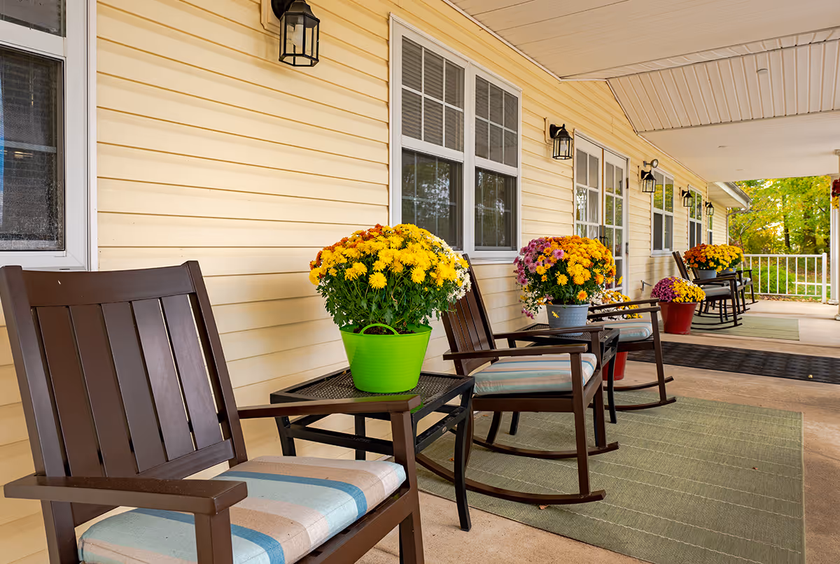 Covered outdoor porch area with several wooden rocking chairs and small tables, each table holding a colorful pot of flowers. The porch has a light yellow siding wall with windows and outdoor lantern-style lights mounted above. Green mats are placed on the concrete floor, and trees with green foliage are visible in the background.