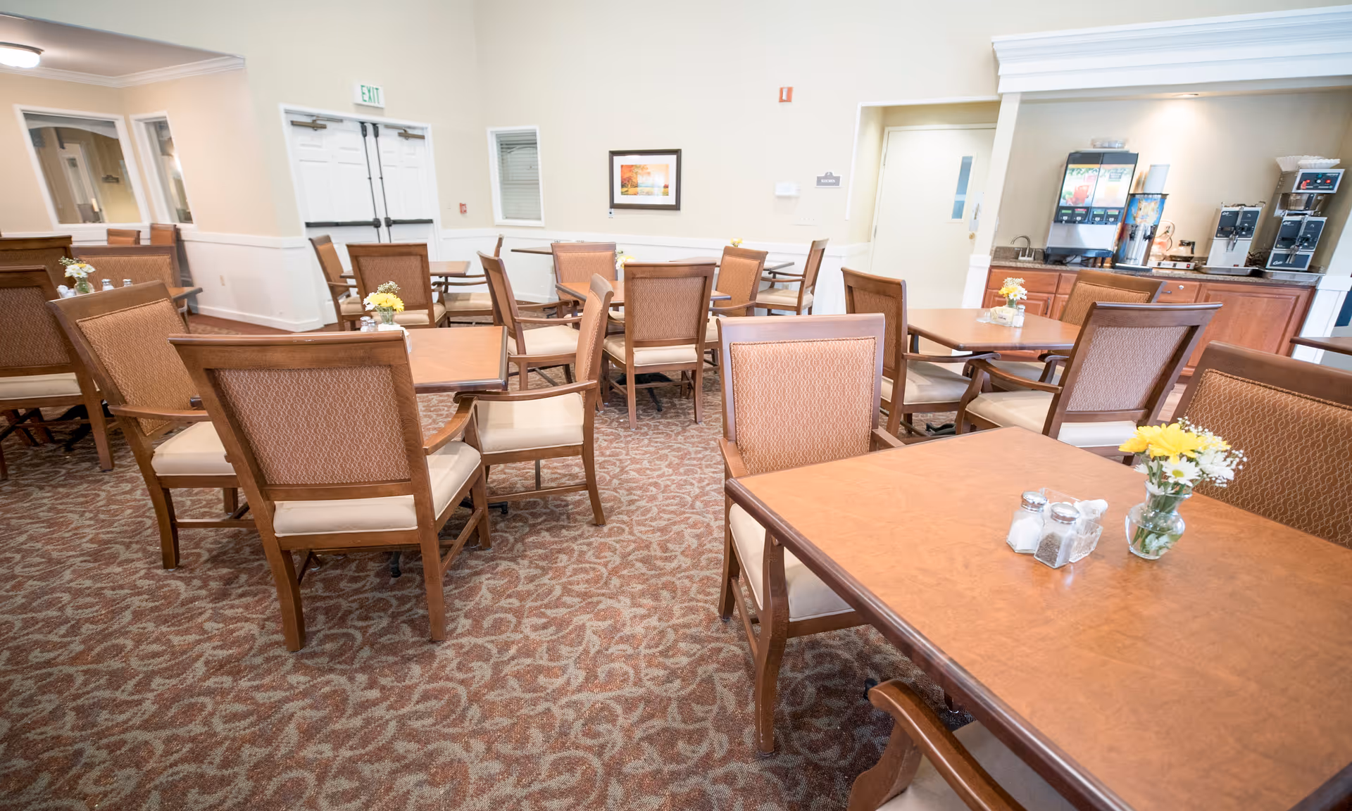 A bright dining room with multiple wooden tables and upholstered chairs, small floral centerpieces, and a beverage station in the back.