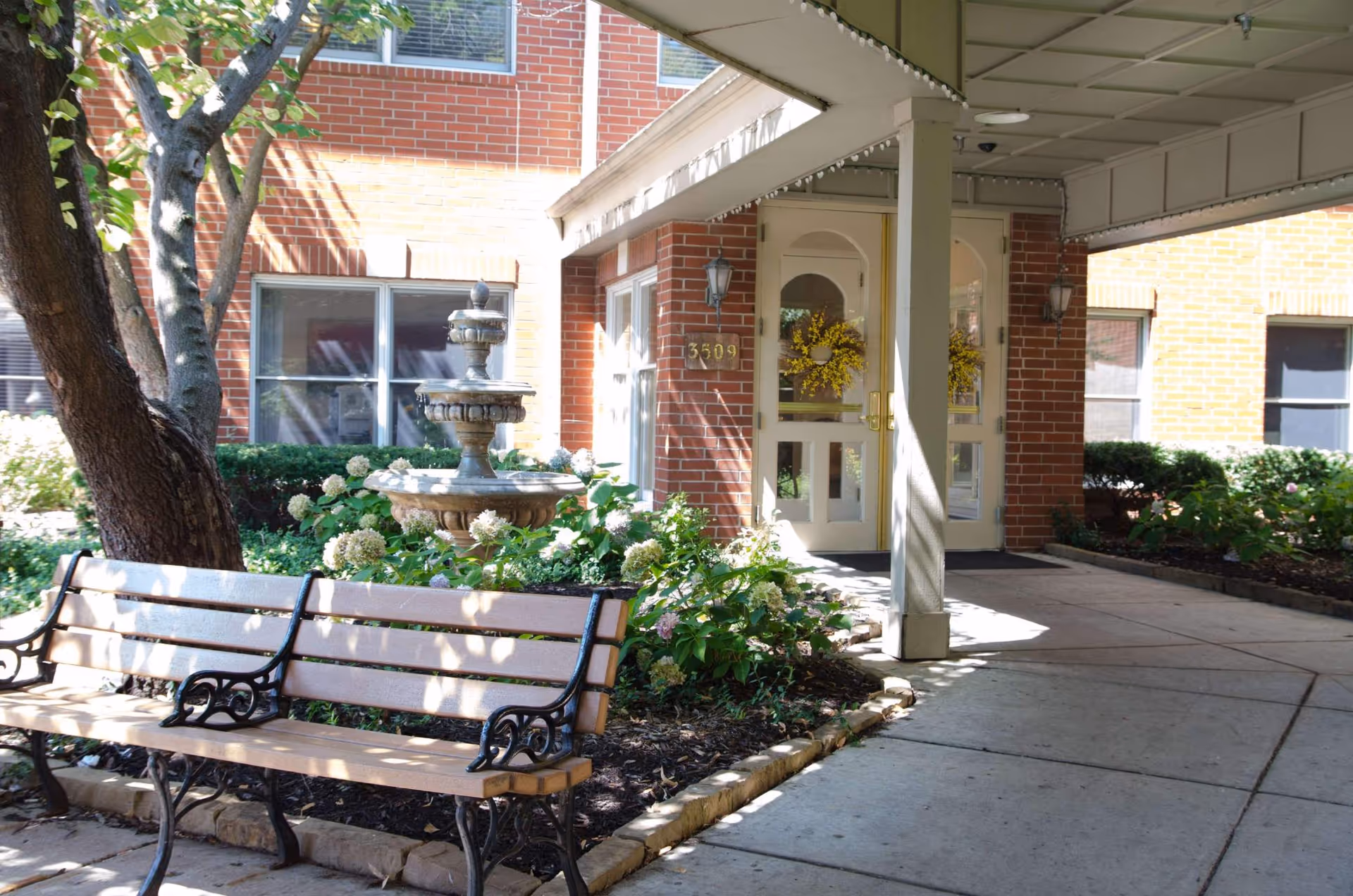 Outdoor entrance area of a brick building with a covered walkway, a double door decorated with a yellow wreath, a stone fountain surrounded by greenery, and two wooden benches with black metal armrests and legs.
