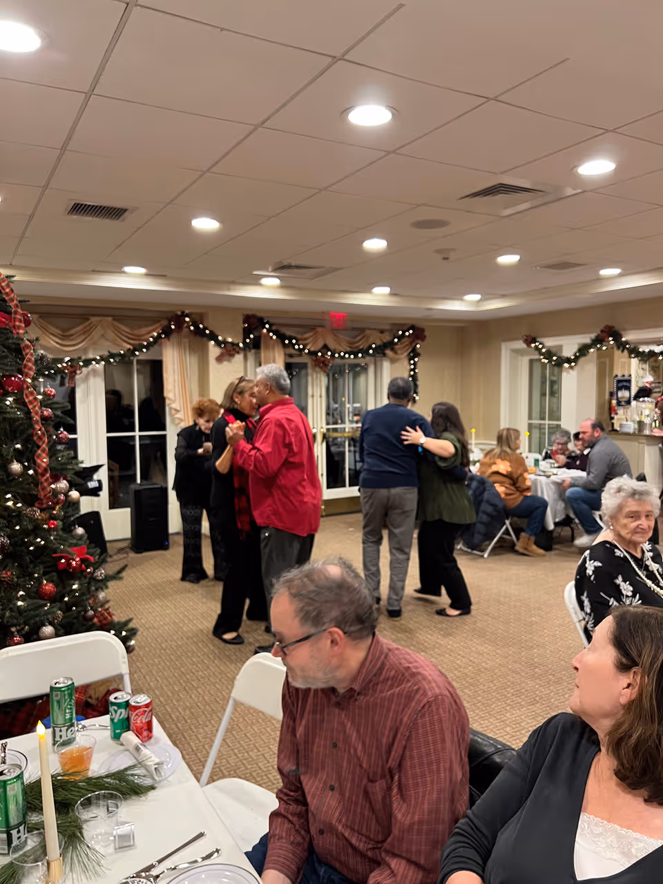 A festive indoor gathering at a senior living facility with several elderly people dancing and sitting at tables. The room is decorated with Christmas garlands and a decorated Christmas tree. People are engaged in conversation and enjoying the event.