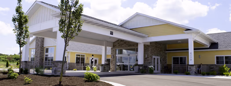 Front exterior view of Vineyard Assisted Living Community building with a covered entrance, yellow siding, stone accents, and landscaped surroundings under a partly cloudy sky.