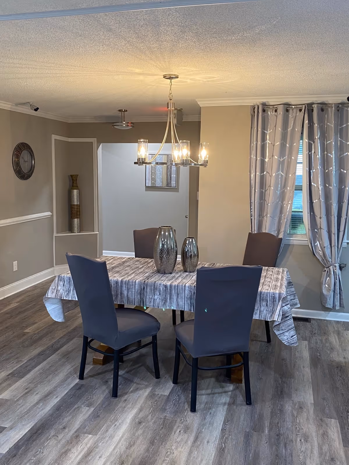 A dining room with a rectangular table covered by a tablecloth that looks like weathered wood. Four chairs with dark gray covers surround the table. Two metallic vases are placed on the table. A modern chandelier with multiple lights hangs above the table. The room has light gray walls, a clock on one wall, a decorative vase in a recessed corner, and a window with gray patterned curtains.