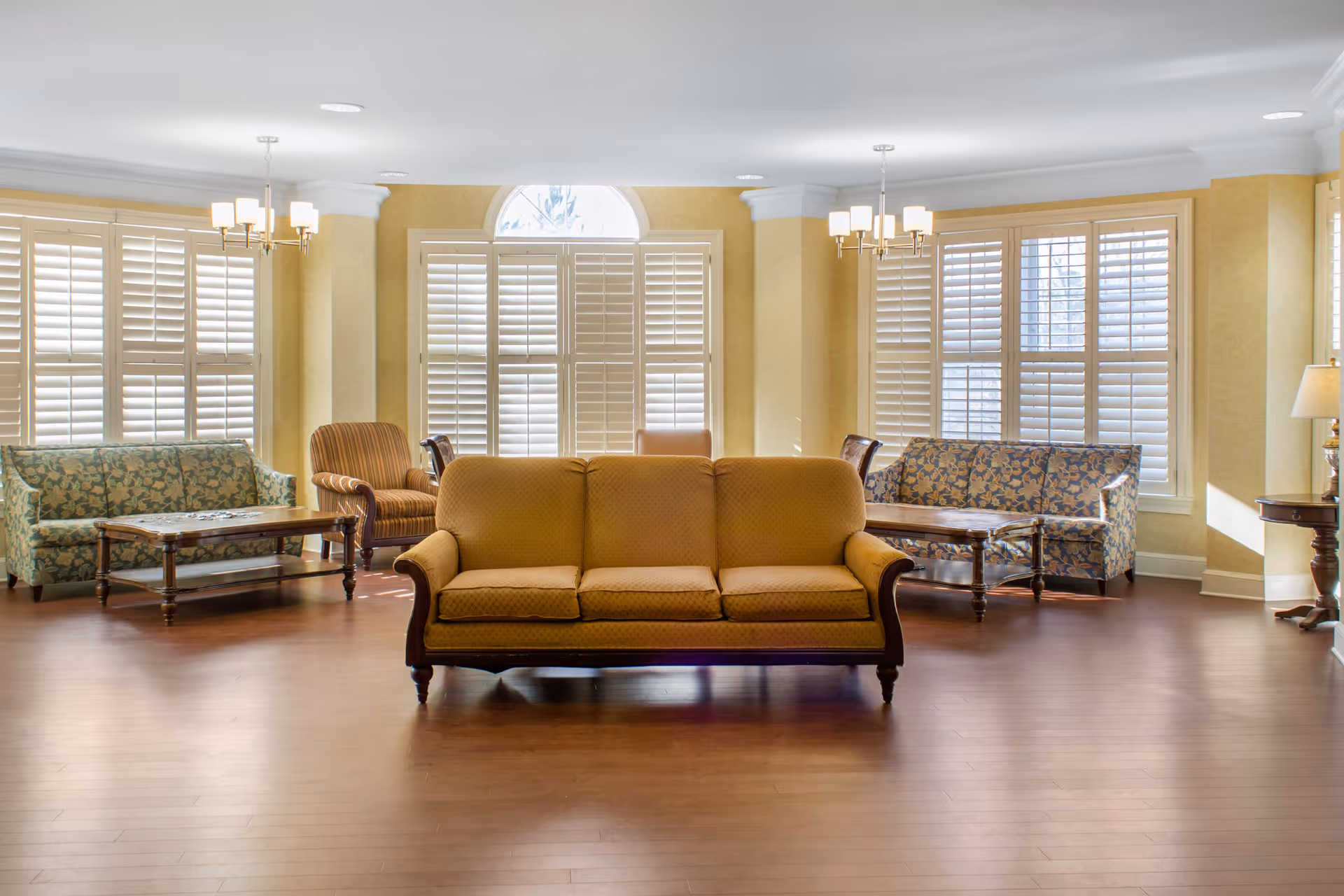 A bright and spacious living room area with multiple sofas and armchairs arranged around wooden coffee tables. The room features large windows with white plantation shutters, yellow walls, and wooden flooring. Two chandeliers hang from the ceiling, and a table lamp is visible on the right side.